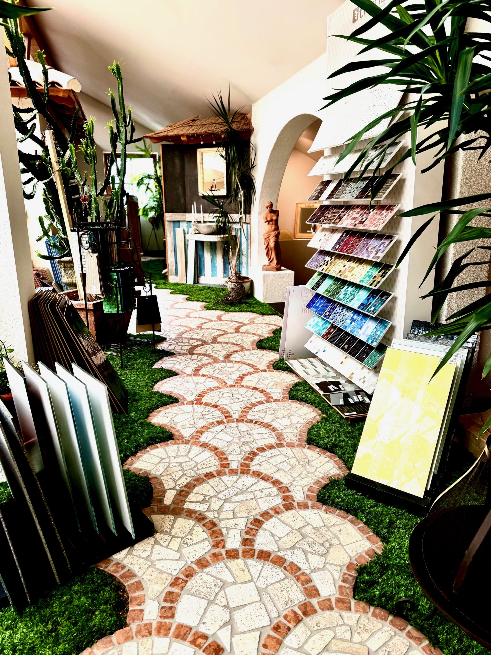 Rechter Gang in der Ausstellung, am Boden sieht man Kunstrasen in der Mitte wurde in Pflasteroprik ein Weg verlegt. links und rechts davon stehen Mosaikmuster in den schönsten Farben.