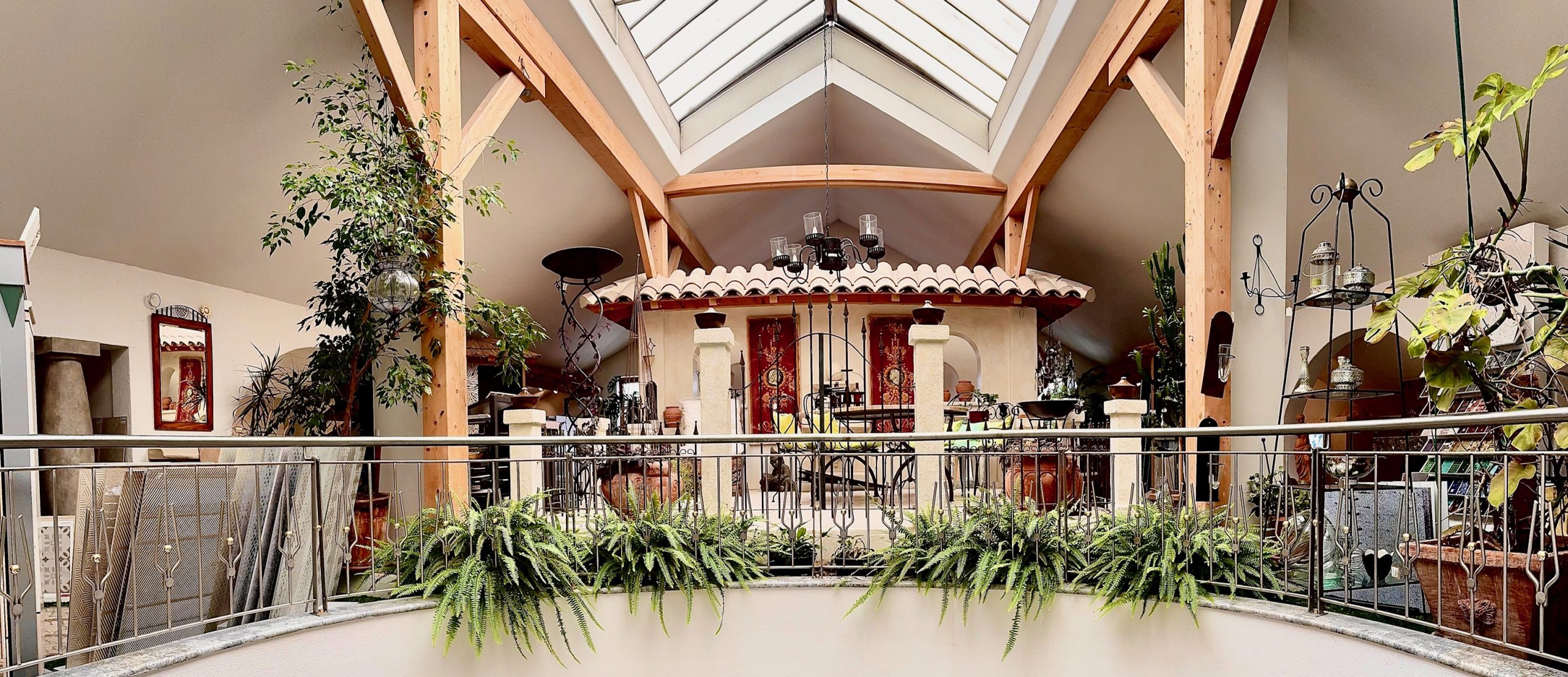 Ausstellung im Dachgeschoss zu sehen ist die sehr grün bepflanzte Galerie mit Holzbalken. In der Mitte sieht man ein Nachgestellten Terrassenbereich im Mediterranen Stil.