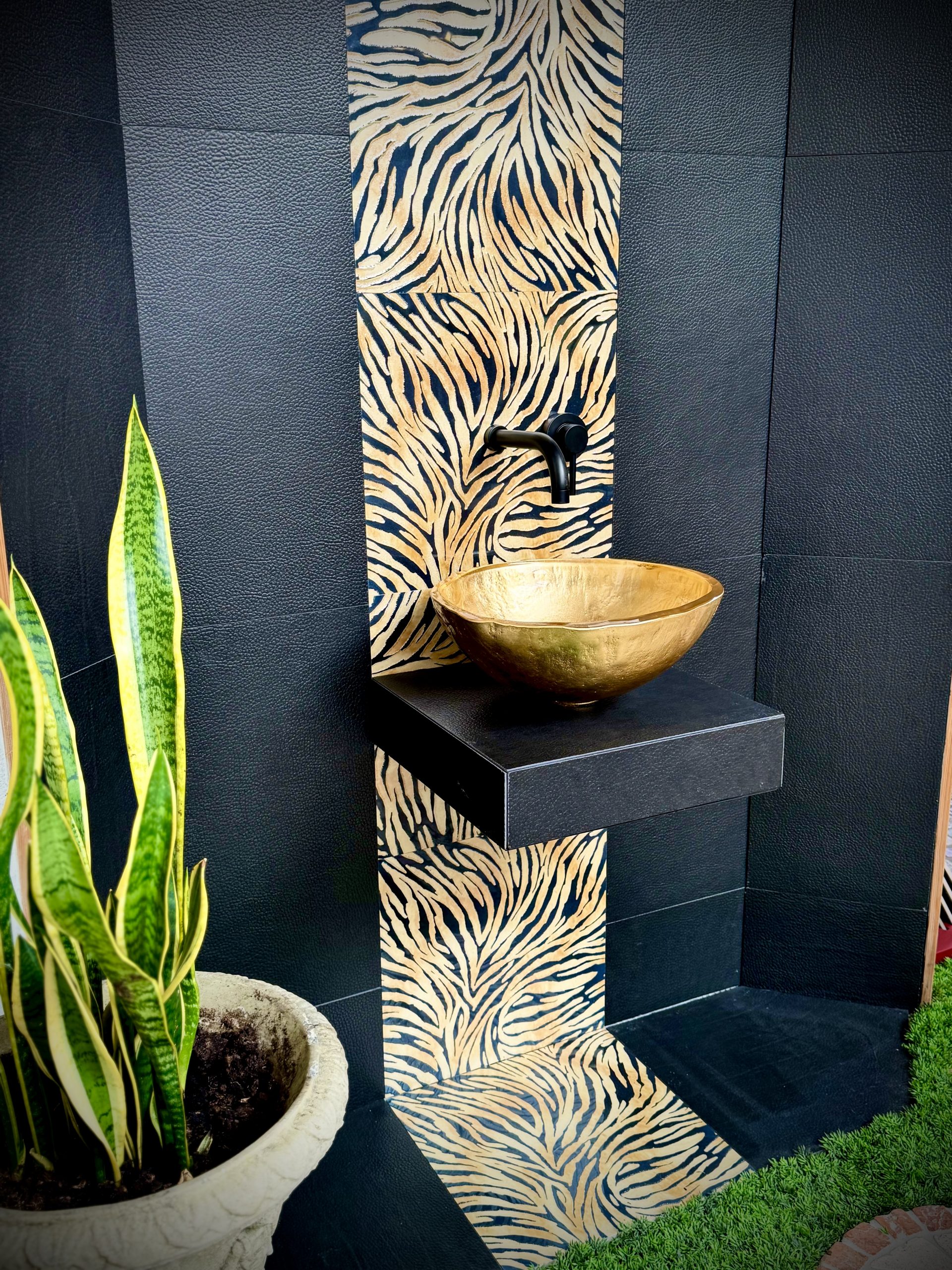 Nische mit schwarzen Wand- und Bodenfliesen. Mittig wurde ein Streifen in goldenen Zebramuster gefliest. Zentral ist ein Goldener Glaswaschtisch der auf einer an der Wand befestigten Vorrichtung steht.