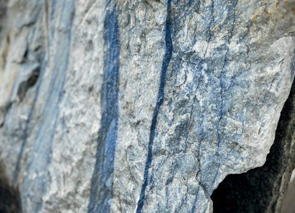 Azul Macaubas Quarzit Ein großer, grob behauener Block aus hellem Naturstein mit markanten, senkrecht verlaufenden blauen Streifen. die Oberfläche ist rau und uneben, mit sichtbaren Bruchkanten und natürlichen Adern. Der Stein zeugt eine Mischung aus hellgrauen und bläulichen Farbtönen. Es handelt sich um einen Quarzit