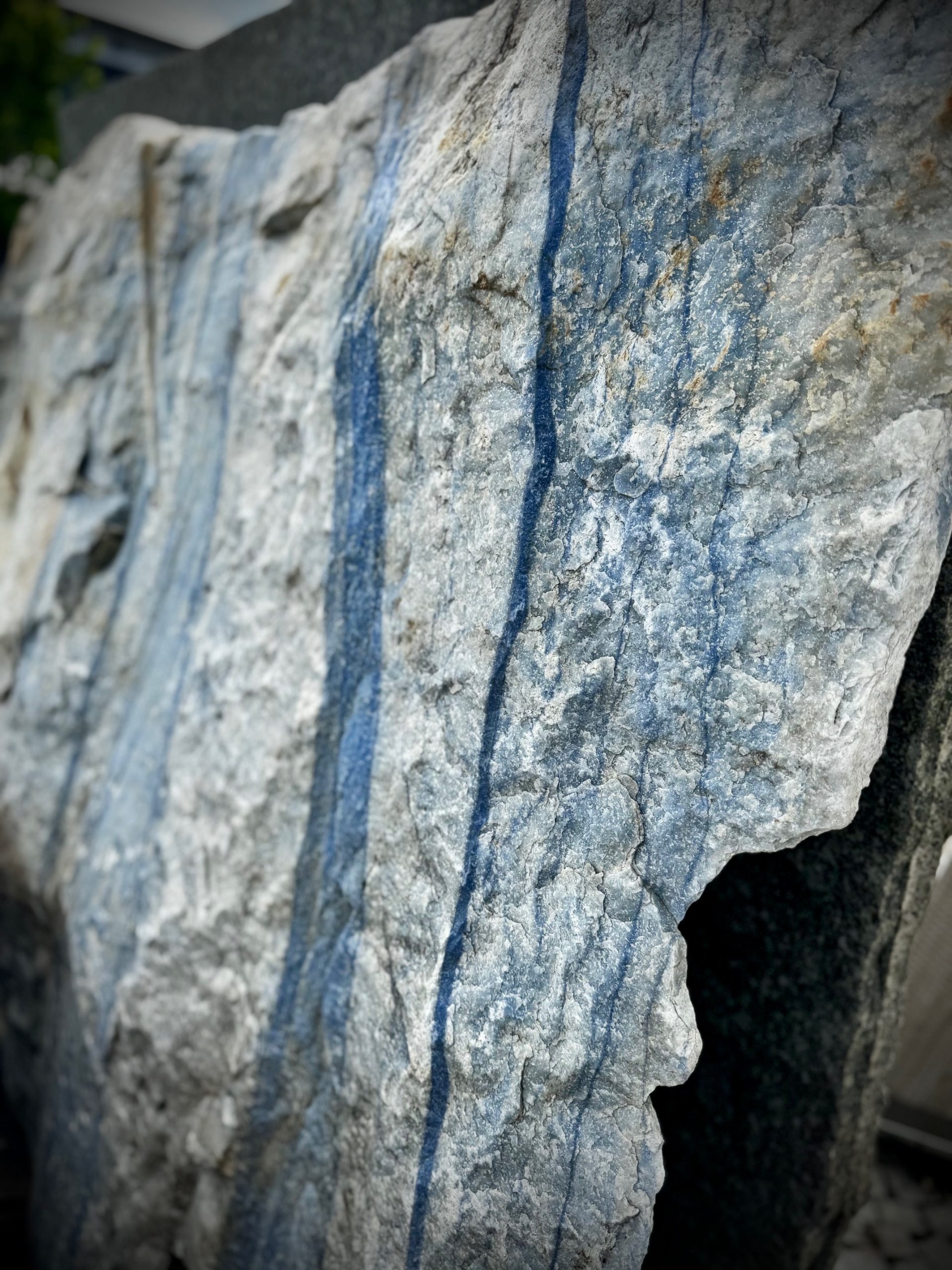 Ein großer, grob behauener Block aus hellem Naturstein mit markanten, senkrecht verlaufenden blauen Streifen. die Oberfläche ist rau und uneben, mit sichtbaren Bruchkanten und natürlichen Adern. Der Stein zeugt eine Mischung aus hellgrauen und bläulichen Farbtönen. Es handelt sich um einen Quarzit