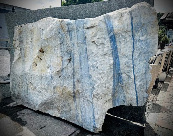 Ein großer, grob behauener Block aus hellem Naturstein mit markanten, senkrecht verlaufenden blauen Streifen. die Oberfläche ist rau und uneben, mit sichtbaren Bruchkanten und natürlichen Adern. Der Stein zeugt eine Mischung aus hellgrauen und bläulichen Farbtönen. Es handelt sich um einen Quarzit